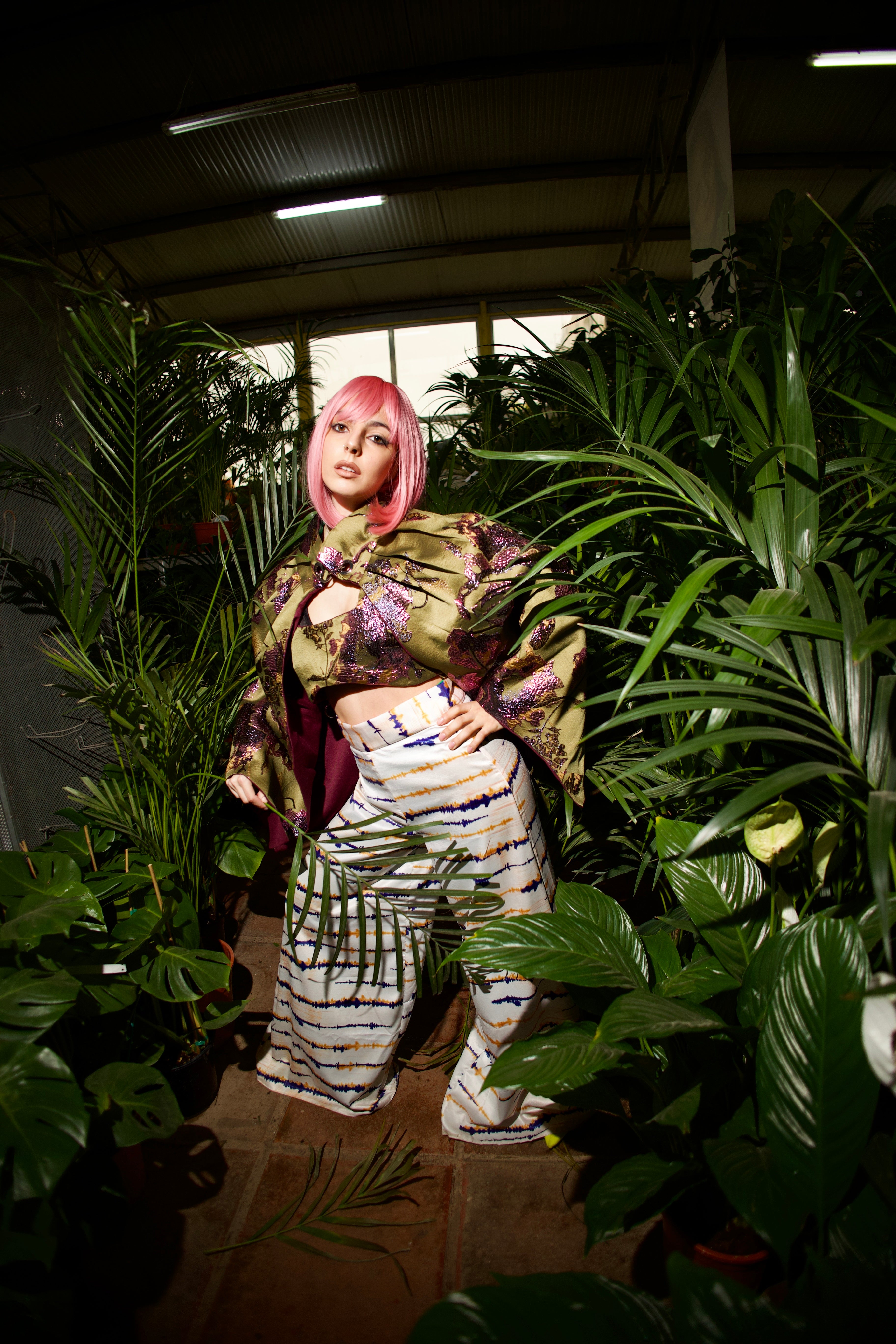 Person with pink hair standing among plants in a dimly lit indoor setting