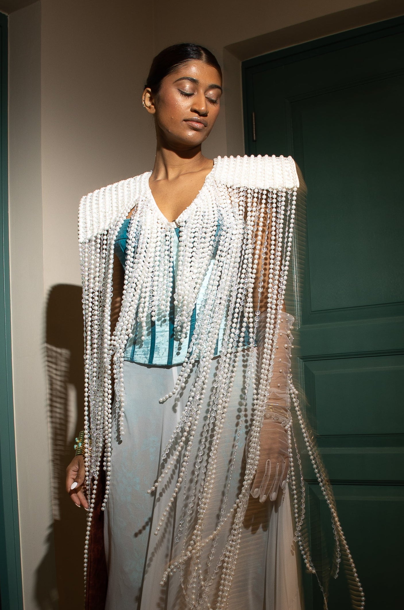 Model wearing a pearl-embellished cape with turquoise corset and sheer white trousers from MVATIS “Mwari’s Gold” haute couture collection, styled with statement accessories, standing between green doors in dramatic lighting.