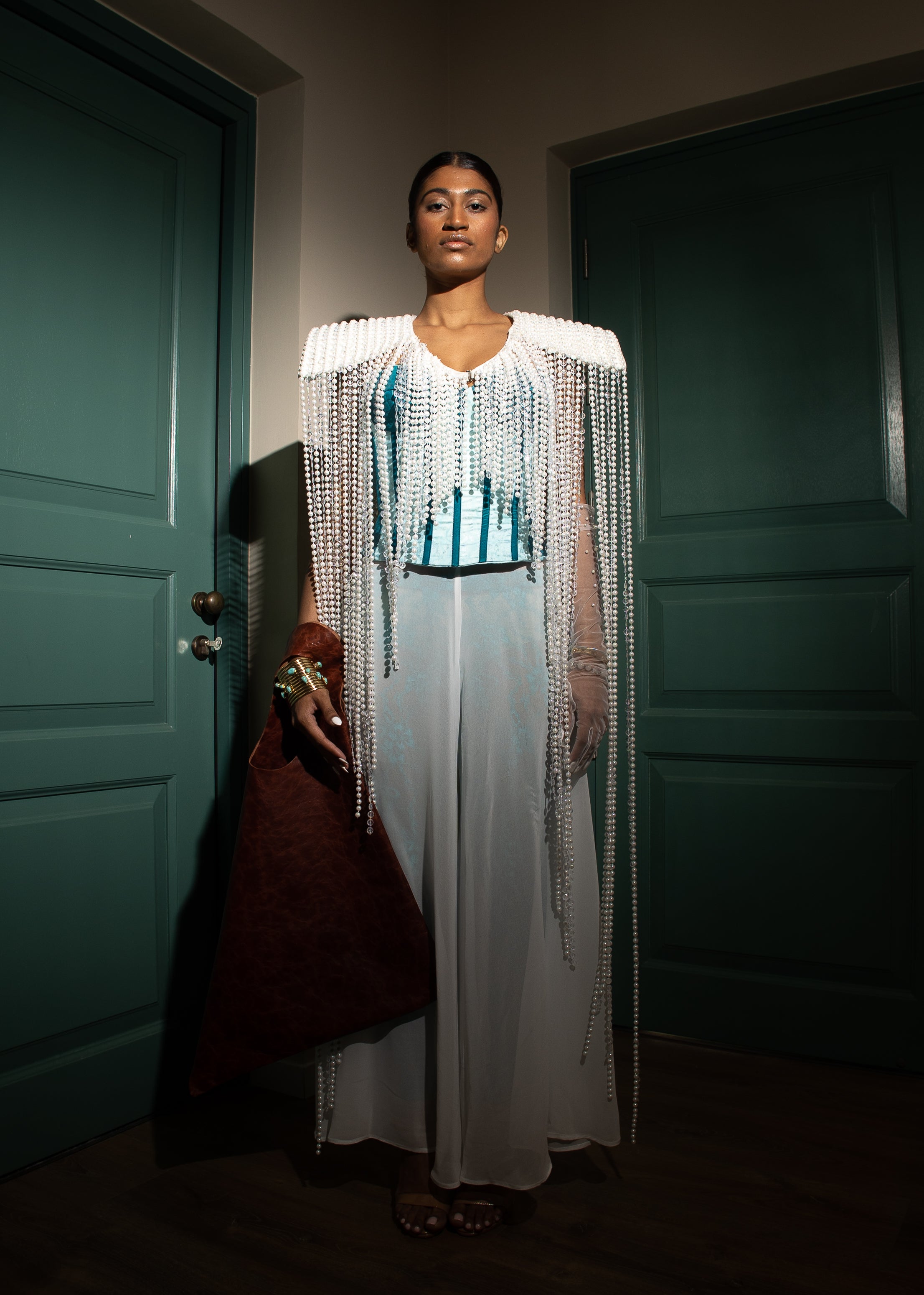 Model wearing a pearl-embellished cape with turquoise corset and sheer white trousers from MVATIS “Mwari’s Gold” haute couture collection, styled with statement accessories, standing between green doors in dramatic lighting.