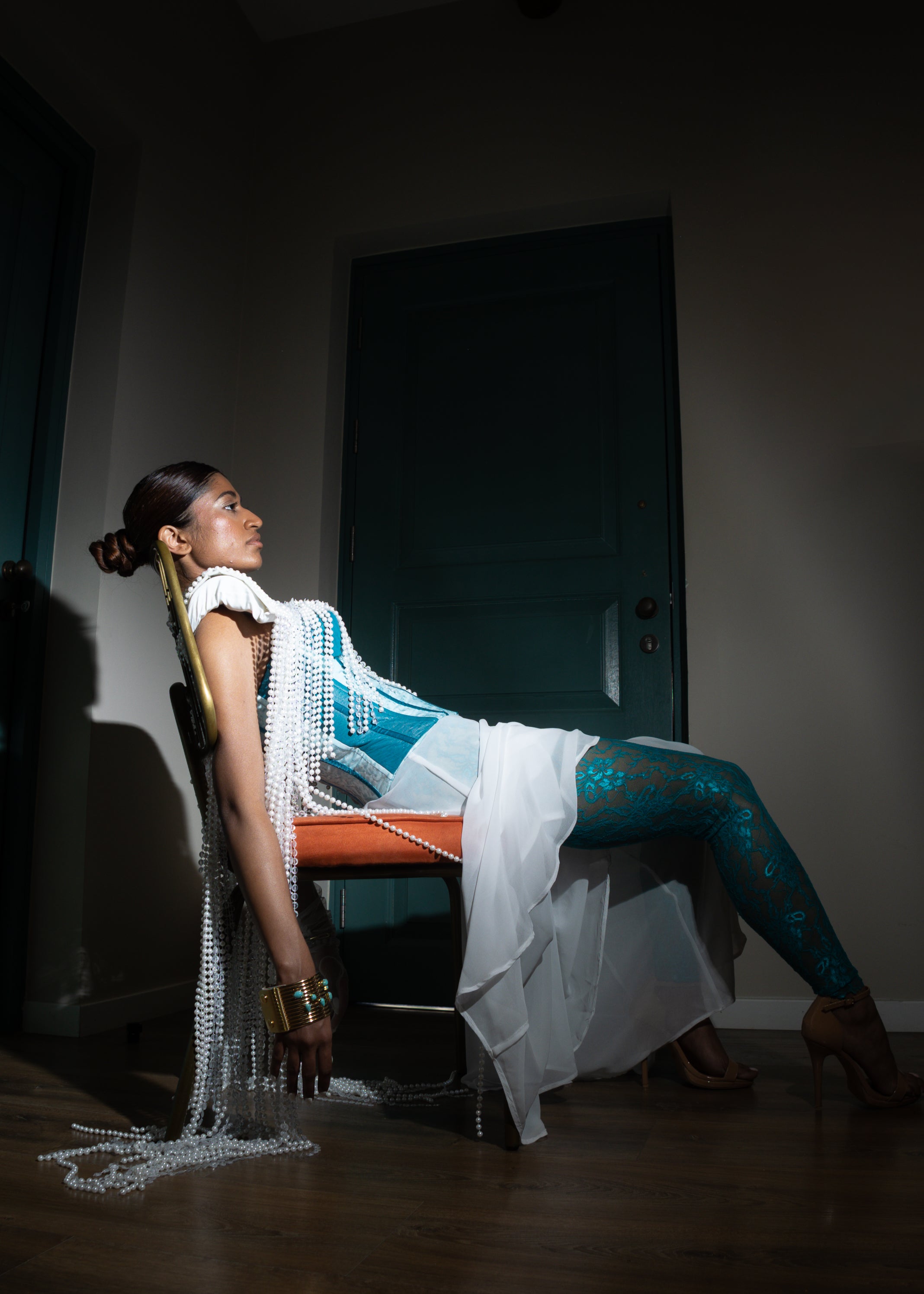 Model wearing a pearl-embellished cape with turquoise corset and sheer white trousers from MVATIS “Mwari’s Gold” haute couture collection, styled with statement accessories, standing between green doors in dramatic lighting.
