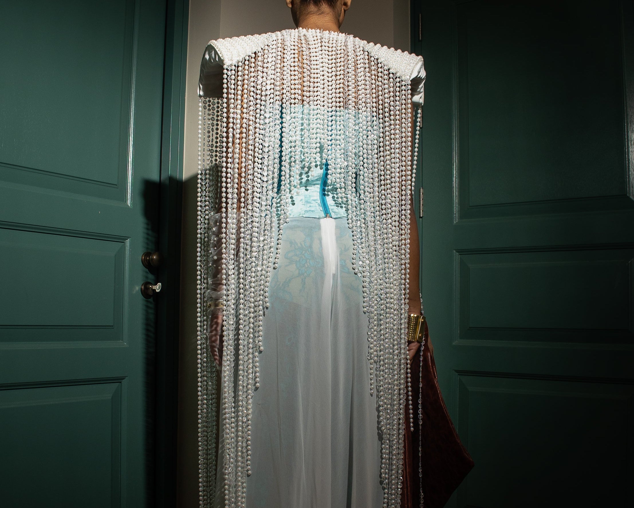 Model wearing a pearl-embellished cape with turquoise corset and sheer white trousers from MVATIS “Mwari’s Gold” haute couture collection, styled with statement accessories, standing between green doors in dramatic lighting.