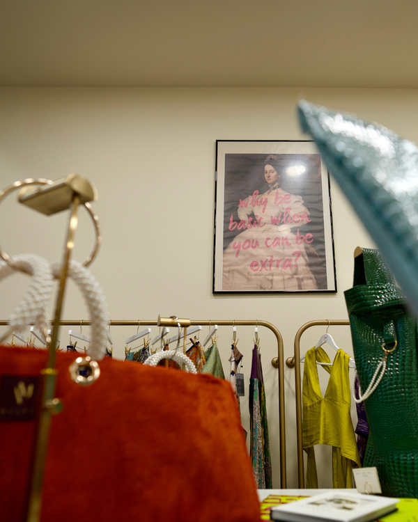 A boutique interior with clothing racks, a framed poster on the wall, and several handbags in the foreground, including a prominent rust-red textured bag on the left and a dark green croc-embossed bag on the right.