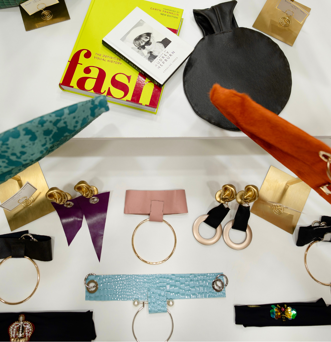 A styled flat-lay display of fashion accessories and a magazine on a white surface. Visible items include a bright yellow 'fashion' magazine, a black round pouch, an orange fabric sleeve, multiple pairs of gold-tone earrings with black and colored accents, several leather or faux-leather ribbon/bow accessories in purple, pink, black, and turquoise, and a green textured handbag corner at the top left.