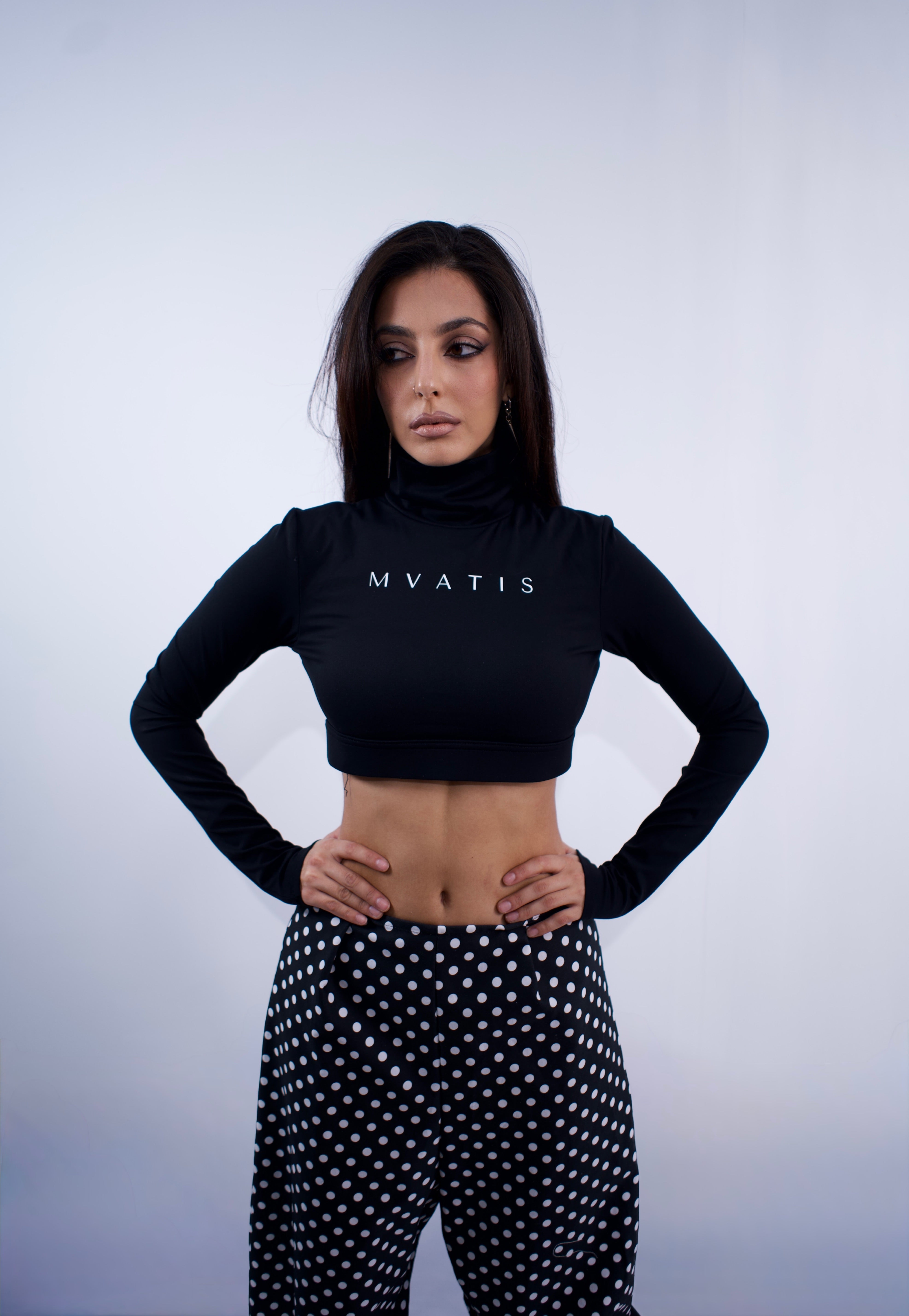 Woman wearing a black crop top with 'MVATIS' branding and polka dot pants on a light gray background