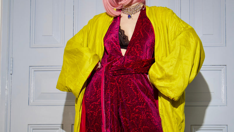 Person wearing a vibrant outfit with a yellow jacket and red dress against a white paneled wall.